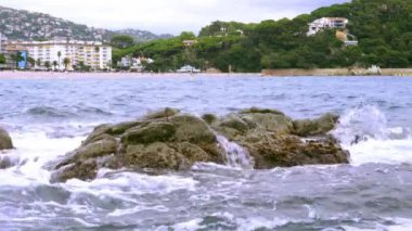 Akşamları deniz dalgaları kumsaldaki kayalara çarpıyor ve arka planda şehir ve orman var.