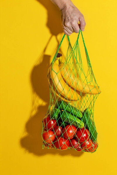 Male hand holding a white mesh bag with vegetables on yellow background