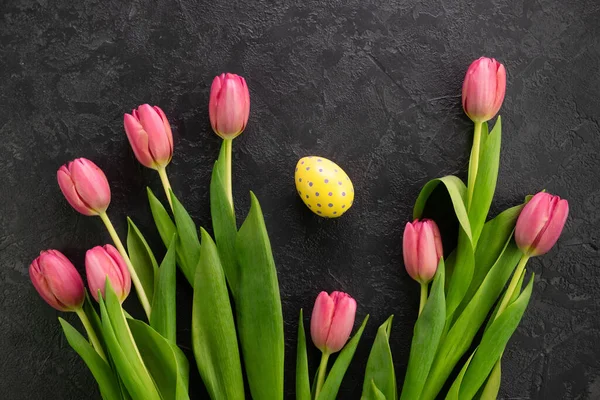 Easter egg and pink tulips on a dark concrete background. Hello spring and easter concept. flowers and colourful egg. Flat lay, top view, Copy space for text