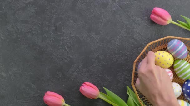 Рука кладет тюльпаны рядом с красочными пасхальными яйцами в корзину на ярком фоне. 4k видео. — стоковое видео