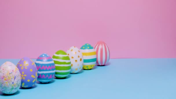 Coloridos huevos de Pascua pintados ruedan y se golpean unos a otros sobre un fondo azul y rosa. — Vídeos de Stock