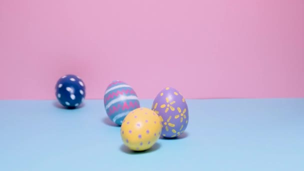 Coloridos huevos de Pascua pintados ruedan y se golpean unos a otros sobre un fondo azul y rosa. — Vídeo de stock