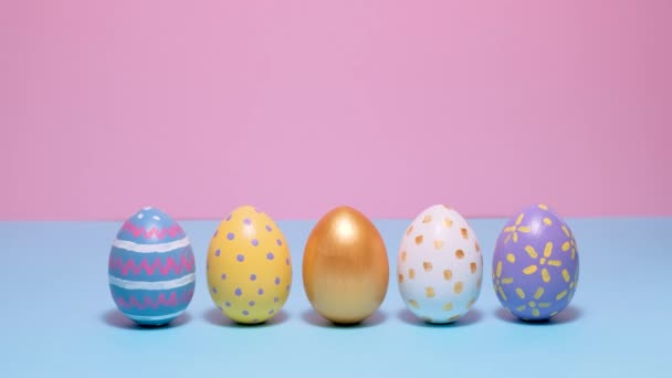 Coloridos huevos de Pascua pintados ruedan y se golpean unos a otros sobre un fondo azul y rosa. — Vídeo de stock