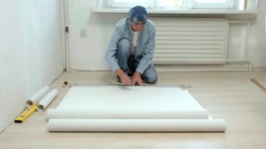 Yerde oturan bir adam bir rulo duvar kağıdı keser. Dairenin tadilatı