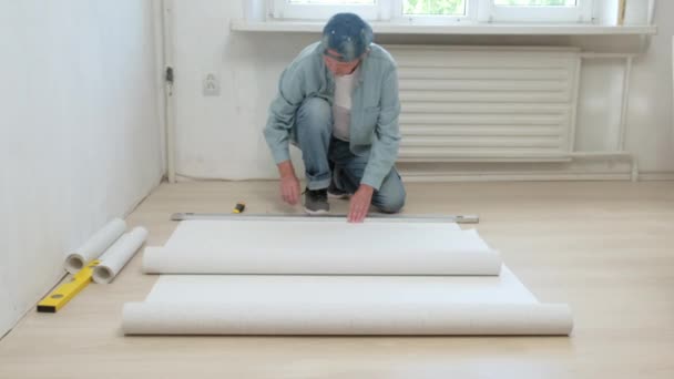 Mężczyzna siedzący na podłodze kroi rolkę tapety. Renowacja mieszkania — Wideo stockowe