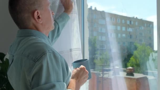 Älterer Mann öffnet Vorhänge und schaut aus dem Fenster, um zu Hause Kaffee zu trinken. — Stockvideo