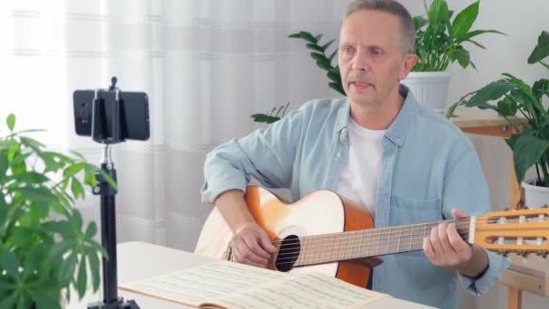 An elderly male blogger teaches guitar playing in an online lesson at home — Stock Video