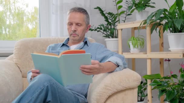 Senior man leest een boek terwijl hij thuis op een fauteuil zit. — Stockvideo