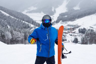 Kışın elinde turuncu snowboard tutan portre ressamı. Mavi ceketli snowboardcu, karlı dağlarda yaşıyor.