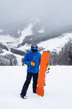Kışın bacağının yanında turuncu snowboard tutan adam. Mavi ceketli snowboardcu, karlı dağlarda yaşıyor.
