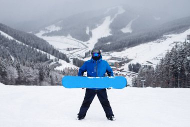 Kışın elinde mavi snowboard tutan adam. Mavi ceketli snowboardcu, karlı dağlarda yaşıyor.