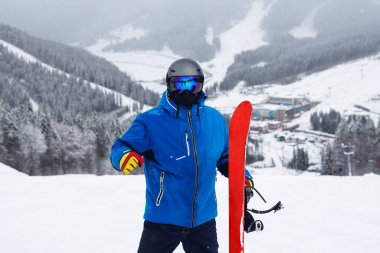Kışın elinde kırmızı snowboard tutan portre ressamı. Mavi ceketli snowboardcu, karlı dağlarda yaşıyor.