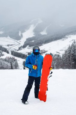 Kışın bacağının yanında kırmızı snowboard tutan adam. Mavi ceketli snowboardcu, karlı dağlarda yaşıyor.