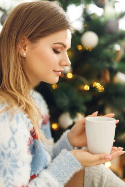 Noel Ağacı 'nın yanındaki evde bir fincan kahve içen bir kızın yan görünüşü. Hayalperest kadın mavi kazaklı, geyik ve gri çoraplı.