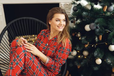 Kızların yan görünüşü şenlikli pijama giyer ve Noel Ağacı 'nın yanında eğlenir. Gülümseyen kadın kırmızı gece elbisesiyle siyah sandalyede oturuyor.