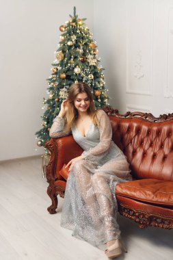 Güzel ve gülümseyen bir kadın Noel Ağacı 'nın yanındaki kahverengi koltukta oturur. Gri elbiseli bir kızla Noel ve Yeni Yıl kavramı aşağı bakıyor