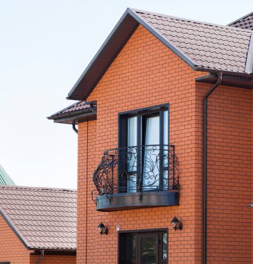 Demir elementlerle işlenmiş güzel bir balkon. Güvenlik kamerası sistemi olan tuğladan bir kır evinin ön cephesi. Özel bir evde güvenlik alarmı. Siyah plastik pencereler ve drenaj sistemi olan metal çatı kiremitleri.