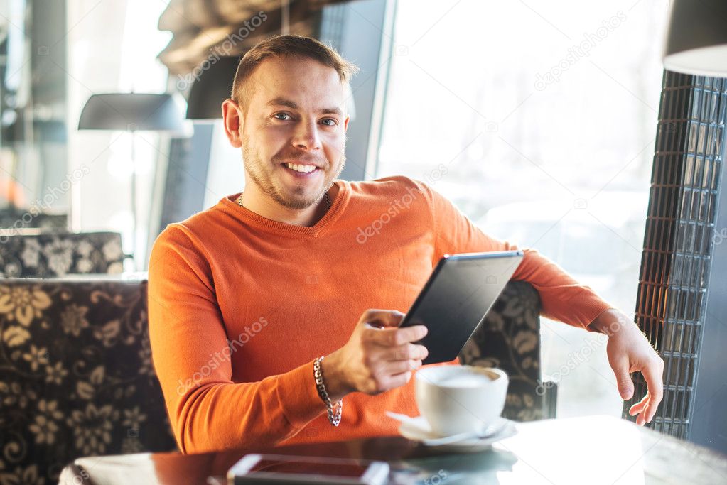 Handsome young man working with tablet, looking at camera, while enjoying coffee in cafe