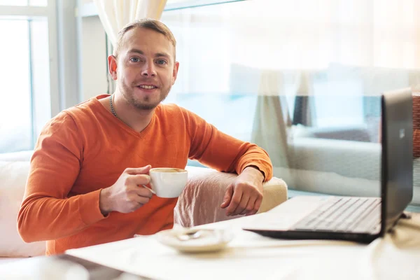 Joven guapo trabajando en el cuaderno, sonriendo, mirando a la cámara, mientras disfruta del café en la cafetería — Foto de Stock