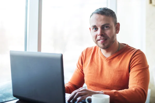 Joven guapo trabajando en el cuaderno, sonriendo, mirando a la cámara, mientras disfruta del café en la cafetería — Foto de Stock