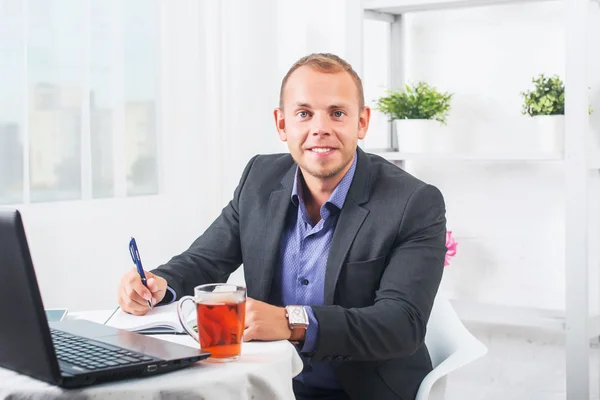 Empresario trabajando en la oficina, sentado en la mesa con un portátil, mirando sonriente . — Foto de Stock