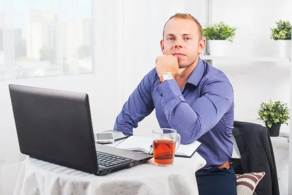 Empresário trabalhando no escritório, sentado à mesa com um laptop, olhando câmera . — Fotografia de Stock