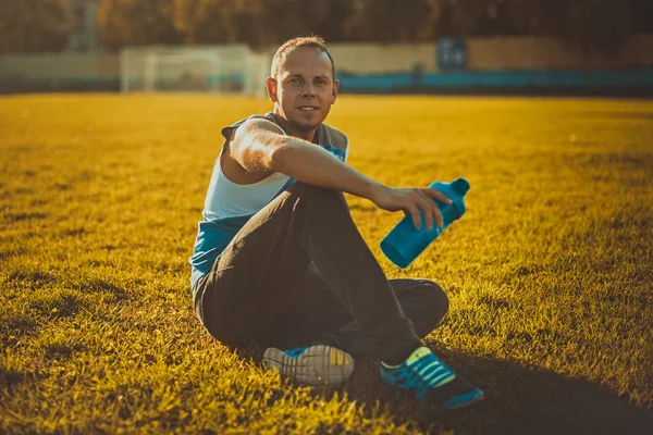 Deportivo un hombre atractivo sentado en la hierba y descansa en el estadio, mantiene el día soleado agitador — Foto de Stock