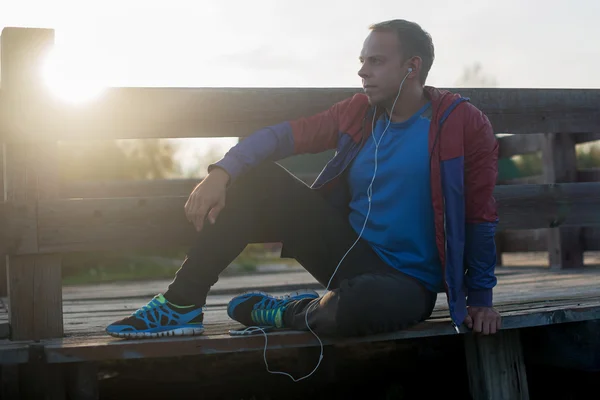 Corredor cansado sentado, relajarse y escuchar música de su teléfono en un muelle de madera, deporte . — Foto de Stock