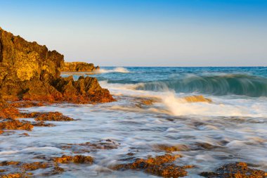 Dalgalar ve su sıçramalarıyla turkuaz deniz ve yaz akşamları kayalık turuncu bir plaj.