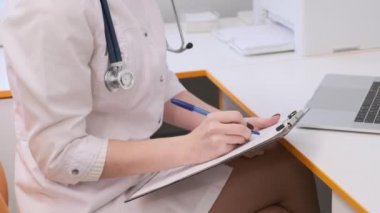 Yakın plan. Beyaz önlüklü genç bir bayan doktor bir hastayla konuşuyor ve araştırma raporunu tamamlıyor.. 