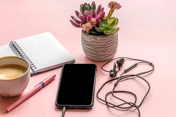 Bir fincan kahve, akıllı telefon, kulaklık, not defteri, kalem ve sulu bitki pembe bir yüzey üzerinde bir demlikte.