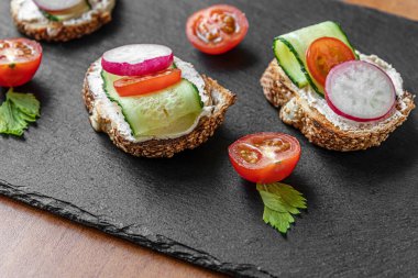 Krem peynirli, salatalıklı, turplu ve domatesli ekmek dilimleri siyah tahtada.