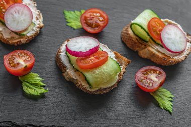 Krem peynirli, salatalıklı, turplu ve domatesli ekmek dilimleri siyah tahtada.