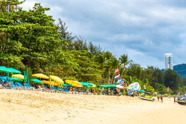 Kathu Phuket Tayland 08. Ekim 2025 Patong Sahili manzaralı gezinti turistleri mavi su dalgaları güneş şemsiyeleri cankurtaran kuleleri dağlar ve Tayland palmiyeleri.