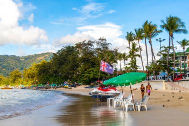 Kathu Phuket Tayland 13 numara. Ekim 2025 Patong Sahili manzaralı gezinti turistleri mavi su dalgaları güneş şemsiyeleri cankurtaran kuleleri dağlar ve Tayland palmiyeleri.
