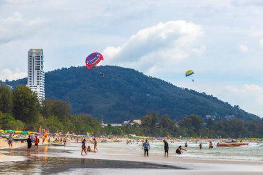 Kathu Phuket Tayland 17 numara. Ekim 2025 Patong Sahili manzaralı gezinti turistleri mavi su dalgaları güneş şemsiyeleri cankurtaran kuleleri dağlar ve Tayland palmiyeleri.