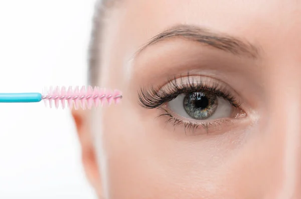 A beautiful light-skinned girl with beautiful eyes combs her extended eyelashes with an eyelash brush. Very close-up soft light isolated white background.