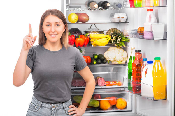 Девушка с длинными волосами в повседневной одежде улыбается и держит указательный палец на фоне открытого холодильника с едой. Концепция диеты или здорового питания.