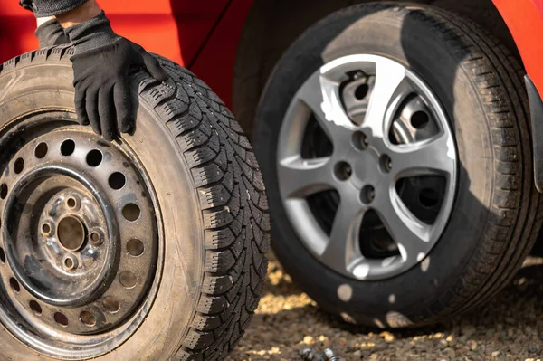 A man rolls a wheel with a studded tire on the background of a red car with a summer tire. The concept of replacing the summer wheel with a winter wheel with your own hands