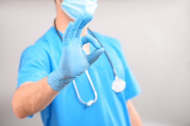 Mavi üniformalı, boynunda steteskop olan tıbbi bir maske takan bir doktor gri arka planda 