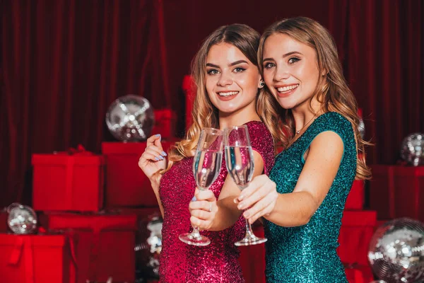 2人の陽気な幸せな女の子は クリスマスを祝う新しい年パーティーを持っています シャンパンとオープンプレゼントを飲む 彼らはセクシーな方法で自分の体を曲げるセクシーなドレスを身に着けている 多くの赤いプレゼントが背中に — ストック写真