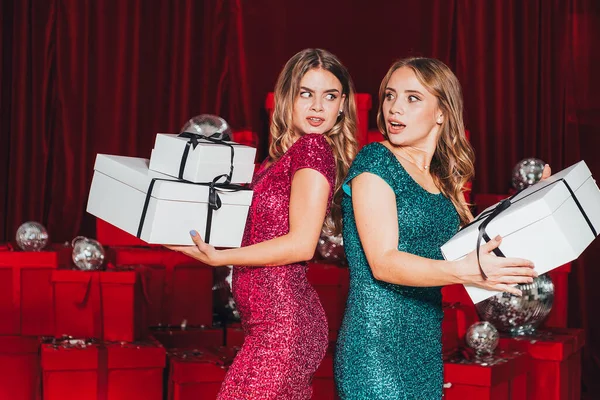 2人の陽気な幸せな女の子は クリスマスを祝う新しい年パーティーを持っています シャンパンとオープンプレゼントを飲む 彼らはセクシーな方法で自分の体を曲げるセクシーなドレスを身に着けている 多くの赤いプレゼントが背中に — ストック写真