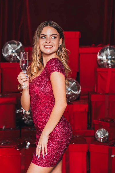 クリスマスを祝う新しい年パーティーを開く陽気な幸せな女の子 シャンパンとオープンプレゼントを飲む 彼女はセクシーな方法で自分の体を曲げるセクシーなドレスを身に着けています 多くの赤いプレゼントが背中に — ストック写真