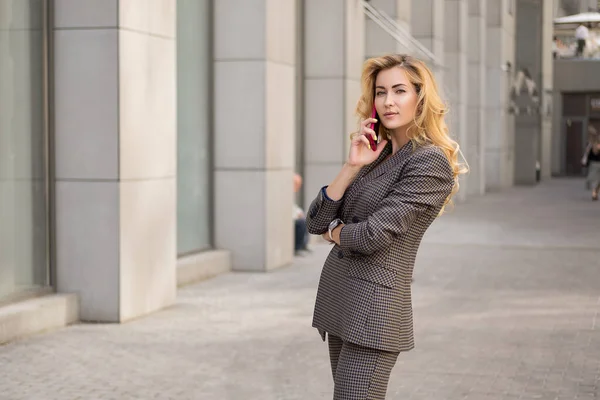 美丽的金发女人穿着灰色经典的智能休闲装站在高科技商业大楼附近 她打了一个电话 案文的篇幅 — 图库照片