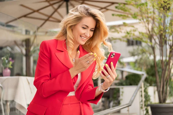 美丽的金发女人穿着红色经典的智能休闲装在高科技商业大楼附近 她在电话里用视频聊天 案文的篇幅 — 图库照片