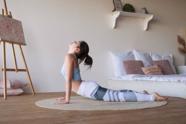 Kadın evde pilates mi yapıyorsun, pilates mi yapıyorsun? Spor giyiniyor ve paspas kullanıyor. Zayıf, esnek ve esmer, beyaz bir kadın. Meditasyon modunda rahatlar ve neşeyle gülümser. Metin için boşluk.