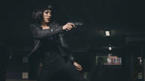 Beautiful brunette sexy spy agent (killer or police) woman in leather jacket and jeans with a gun in her hand running after someone, to catch him on parking.