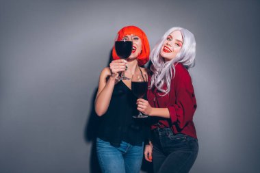 Tarafsız gri arka planda kot ve bluz giymiş kırmızı ve gri renkli peruklar takmış iki tatlı sarhoş kız. Onlar mutlu bir şekilde şarap içiyorlar. Haşin moda aydınlatmasıyla gülümseyen hayat tarzı portresi