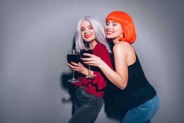 Две милые пьяные девушки в красных и серых париках, одетые в джинсы и блузки на нейтральном сером фоне. Они с радостью пьют вино. Улыбающийся портрет с резким освещением моды