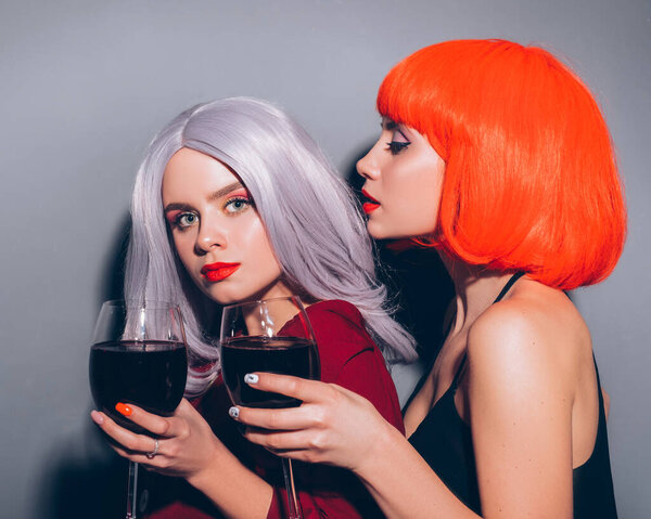 Две милые пьяные девушки в красных и серых париках, одетые в джинсы и блузки на нейтральном сером фоне. Они с радостью пьют вино. Улыбающийся портрет с резким освещением моды
